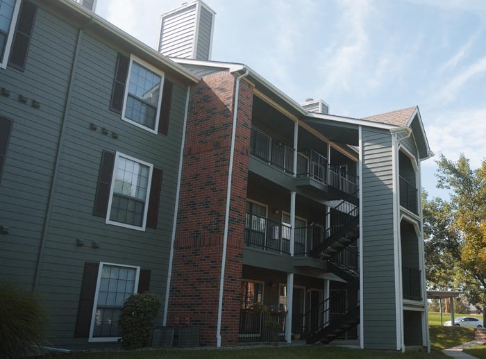 Three-story apartment building with sage green siding, brick accents, and dark shutters